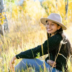 A smiling adolescent girl with long dark hair sitting in a golden autumn field of tall grass. She's wearing a beige wide-brimmed hat, a dark sweater, and jeans. The background shows blurred yellow and gold fall foliage creating a warm, peaceful atmosphere. The girl appears relaxed and content, with sunlight illuminating her genuine smile.