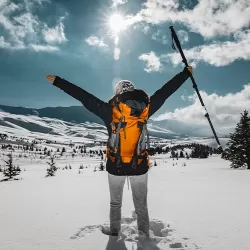 A hiker standing in deep snow with arms outstretched in triumph, viewed from behind. The person is wearing a bright orange backpack and holding a trekking pole, facing a vast snow-covered mountain landscape under a sunny blue sky with scattered clouds. The image conveys a sense of accomplishment, freedom, and the beauty of winter wilderness.