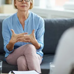 A professional woman in a light blue blouse sits on a gray couch during what appears to be a therapy session. She has short blonde hair and glasses, and is gesturing with her hands while engaged in conversation. The image shows her from the chest up, with her crossed legs visible wearing light pink pants. Papers and a glass of water can be seen in the foreground, suggesting a therapeutic or counseling environment.